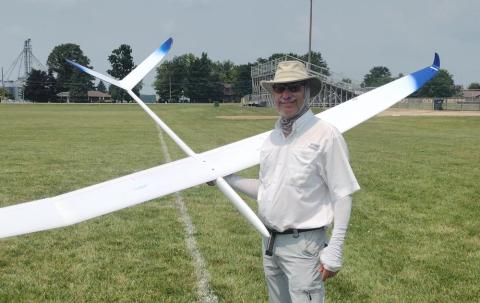 Man in hat holding a white model glider on a grassy field.