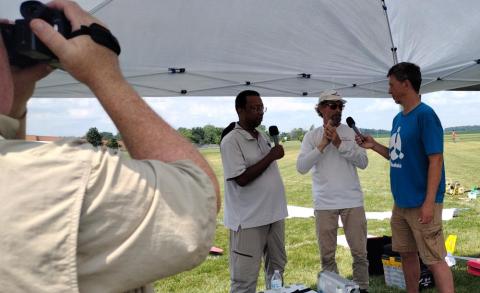 Three men speaking under a tent, with a person filming them, outdoors.