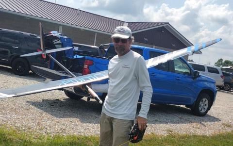 Man holding a large model airplane, standing next to parked trucks on a sunny day.