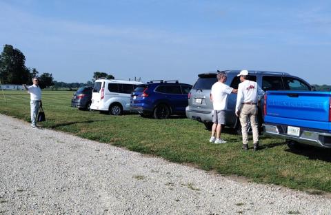 Cars parked on grass, with people standing nearby under a clear blue sky.