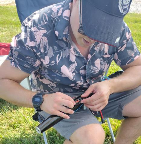 Man seated on grass in floral shirt examines equipment.
