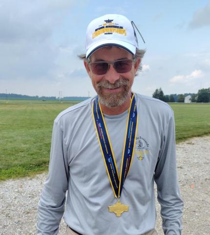 Man smiling outdoors, wearing a cap, sunglasses, and a medal, standing on a grassy field.