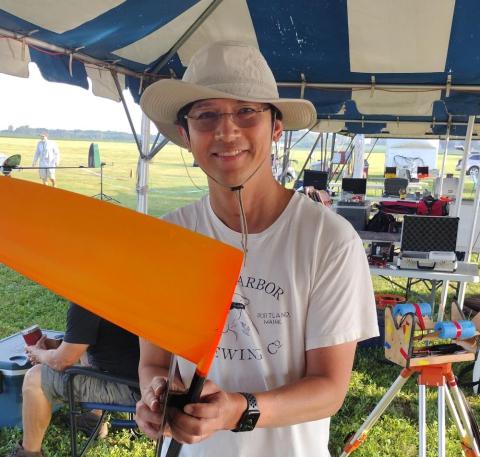 Man wearing a hat holds an orange rocket outdoors under a tent.
