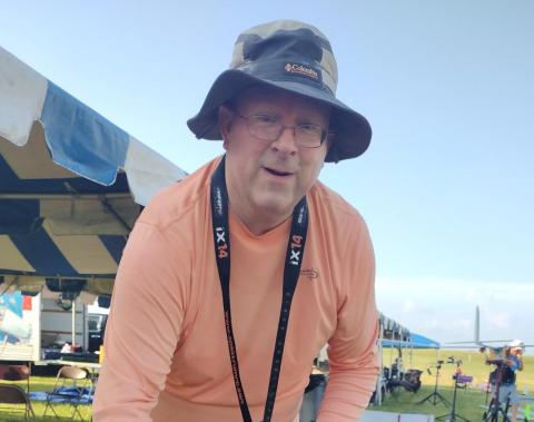 Man in a hat and orange shirt outdoors under a tent.