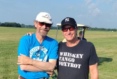 Two smiling men wearing casual clothes and sunglasses, standing in a grassy field.