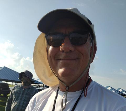 Man in sunglasses and sun hat smiling outdoors under a clear sky.