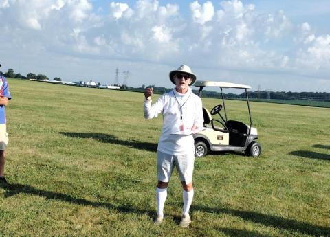 Man in white attire stands on grassy field near golf cart.