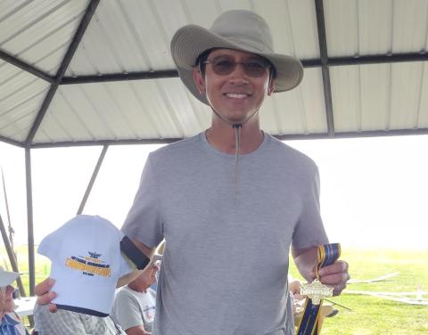 Man in hat holding a white cap and gold medal, smiling under a pavilion.
