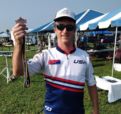 Man in USA cycling jersey holding a medal, outdoors under blue tents.