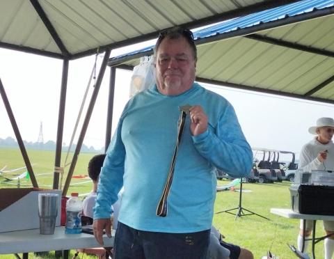 Man in a light blue shirt holding a medal under a canopy at an outdoor event.