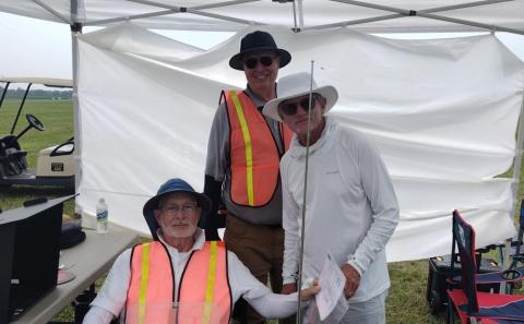 Three men in hats and vests under a white canopy at an outdoor event.