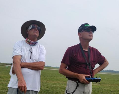 Two men observe the sky in a grassy field, one holding a controller.