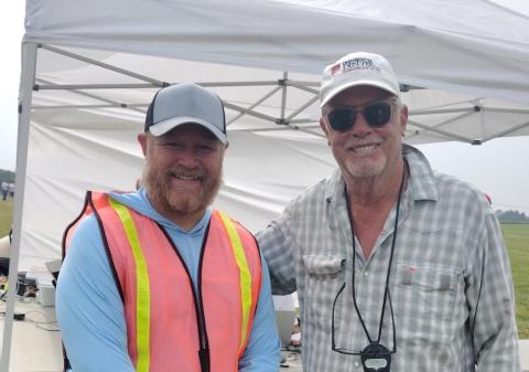 Two smiling men under a tent, one in a safety vest.