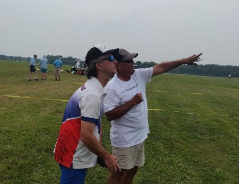 Two men outdoors looking into the distance, one pointing, with a grassy field background.