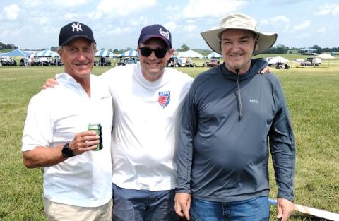 Three men smiling outdoors on a sunny day, wearing casual hats and shirts.