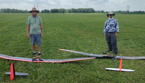 Two people stand on grass near remote-controlled gliders with orange accents.