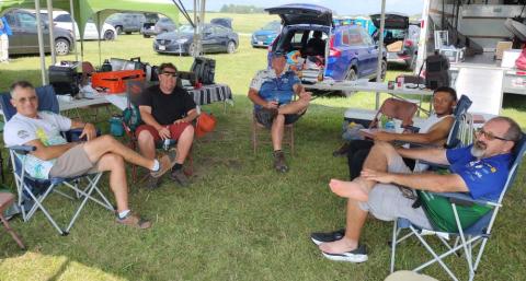 Five people sitting on camping chairs in a grassy area, smiling under shade.