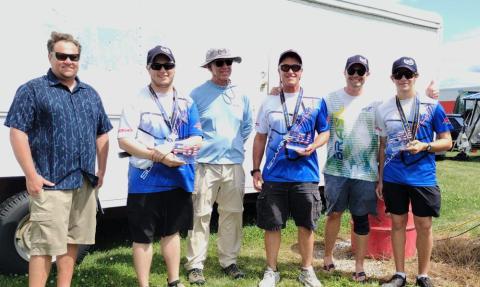 Six people stand together outdoors, some holding small trophies, smiling at the camera.