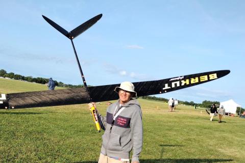 Man in a field holding a large model glider, smiling under a sunny sky.