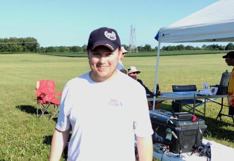 Man smiling in a grassy field near a white canopy, with equipment and chairs visible.