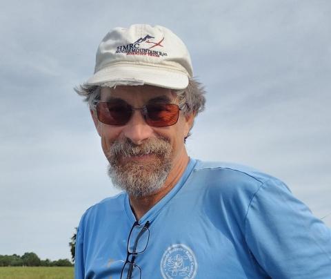 Man in sunglasses and a cap, wearing a light blue shirt, outdoors with cloudy sky.