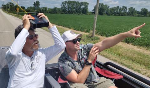Two men sitting in an open vehicle, one holding a black device, the other pointing ahead.