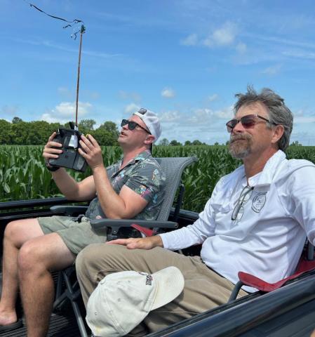 Two men sitting in a truck bed, flying a remote-controlled drone over a field.