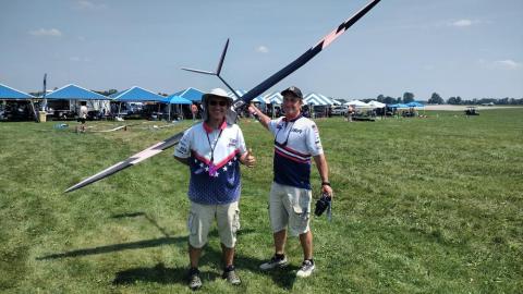 Two men smiling, holding a large remote-controlled glider on a grassy field.