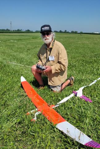 Man kneeling on grass field with orange and white model airplane, sunny day.