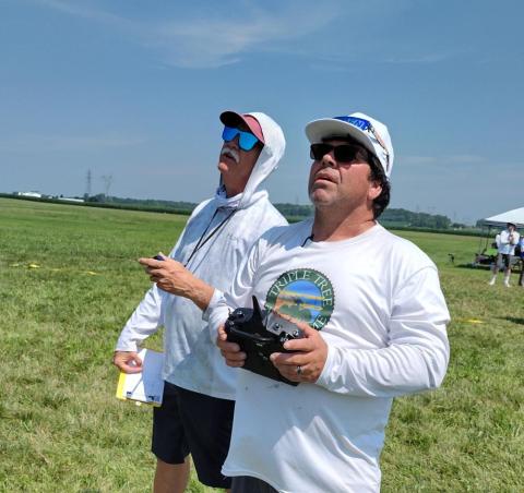 Two men outdoors flying drones with remote controls, under a blue sky.