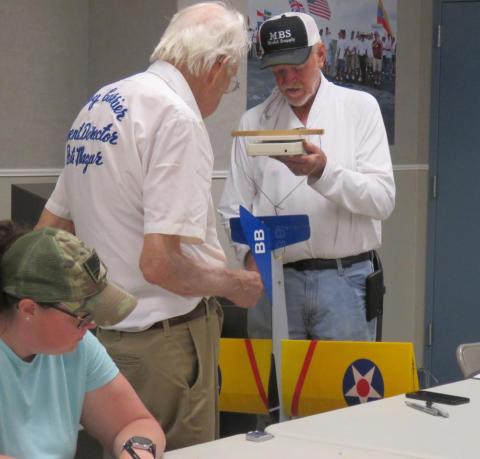 Two men examine a small model rocket indoors, one seated woman in foreground.