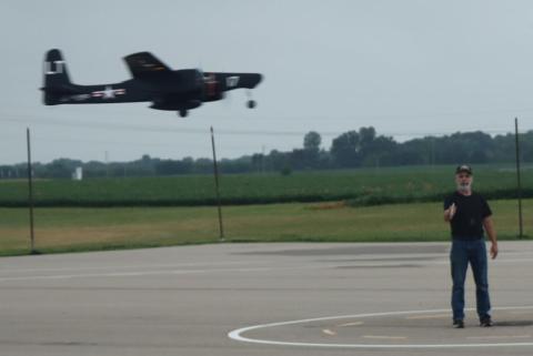 Man standing on tarmac as a vintage military plane flies by.