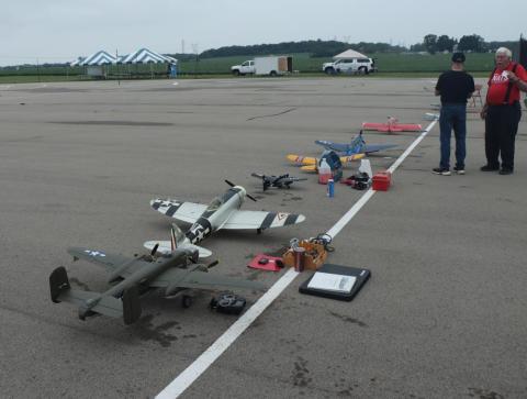 RC planes lined up on an airstrip with two people nearby.