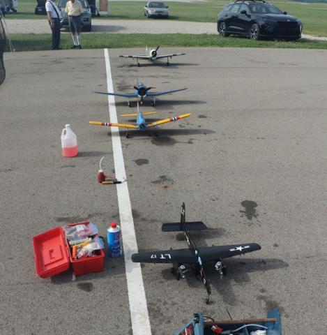 Row of model airplanes on asphalt, tools and supplies nearby, cars and grass in the background.