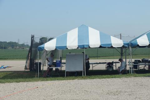 White and blue striped tent on gravel near a grassy field, with people seated and standing inside.