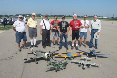 Group of men standing behind model airplanes on an outdoor tarmac.