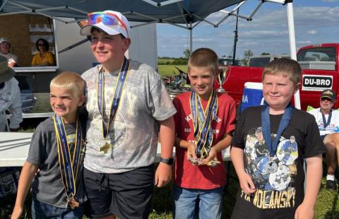 Four smiling boys wearing medals in an outdoor setting under a sunny sky.
