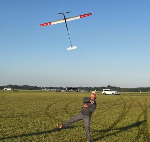 A man launches a glider in a vast grassy field under a clear blue sky.