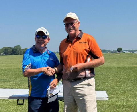 Two men smiling and shaking hands in a grassy field under a clear blue sky.