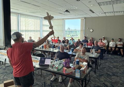 Person in a red shirt holding a cross, speaking to a group seated in a well-lit classroom.