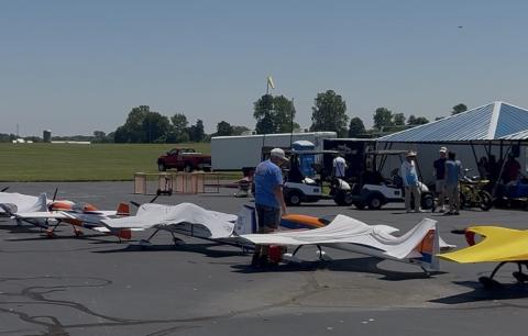 Small model airplanes lined up on an airfield with people nearby under a tent.