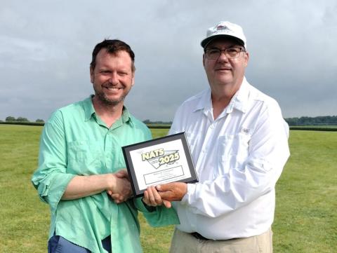 Two men smiling, holding an award plaque outdoors.