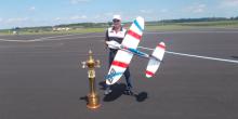 Man with model airplane, standing beside large trophy on airfield.