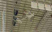Model airplane flying indoors near a high ceiling with lights and signs.