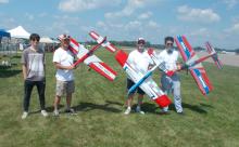 Group of four men outdoors holding red and blue model airplanes.