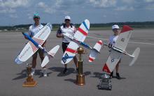 Three people outdoors with large model airplanes and trophies.