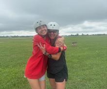 Two people in helmets hugging on a grassy field, smiling under a cloudy sky.