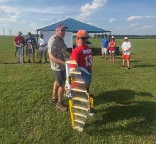 Two men shaking hands at a model airplane event with spectators nearby.