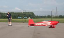 Red model plane flying low near a person on an airfield.