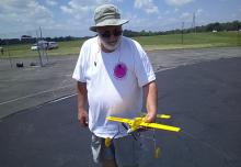 Man holding a yellow model airplane on a sunny day at an airfield.